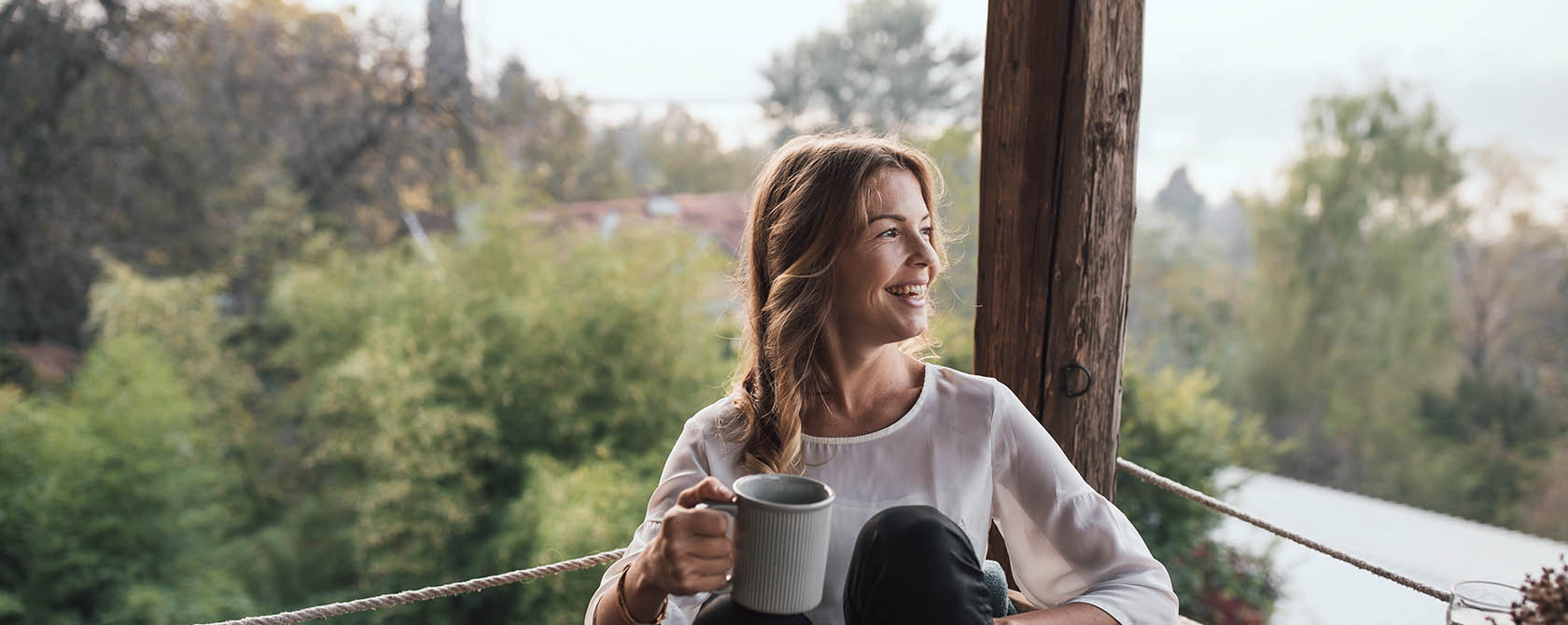 Woman smiling, drinking out of a mug on a balcony. She looks off to the distance.